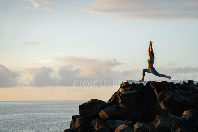 Side view of anonymous female practicing yoga with raised arms on boulders against ocean under cloudy sky at sundown — Stock Photo