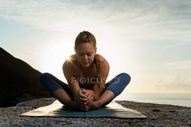 Piano terra di giovane femmina con gli occhi chiusi che si estende gambe e schiena durante la pratica dello yoga sulla costa oceanica sotto il sole — Foto stock