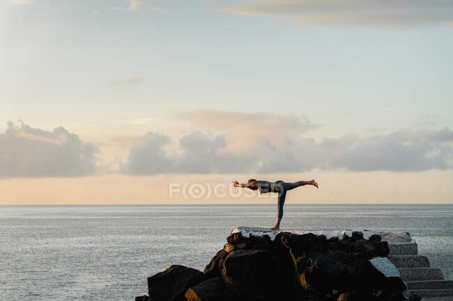Боковой вид неузнаваемой женщины, стоящей в позе Вирабхадрасаны, практикующей йогу на валунах против моря в сумерках — стоковое фото