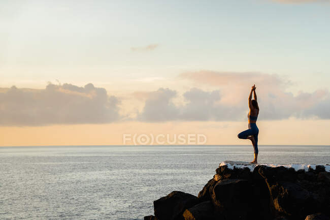 Vue latérale de la femelle méconnaissable debout à Vrksasana pose tout en pratiquant le yoga sur des rochers contre l'océan ondulé dans la soirée — Photo de stock