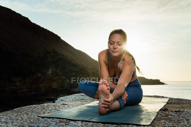 Ground level of young female stretching legs and back while practicing yoga on ocean coast in sunshine — Stock Photo