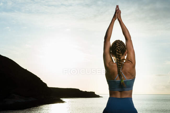 Rückansicht einer nicht wiederzuerkennenden Frau, die in Vrksasana-Pose steht, während sie abends Yoga auf Felsbrocken gegen den wogenden Ozean praktiziert — Stockfoto