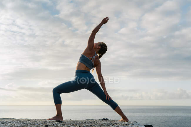 Vista posteriore della femmina in abbigliamento sportivo praticare yoga con braccio sollevato sulla riva dell'oceano sotto cielo nuvoloso — Foto stock