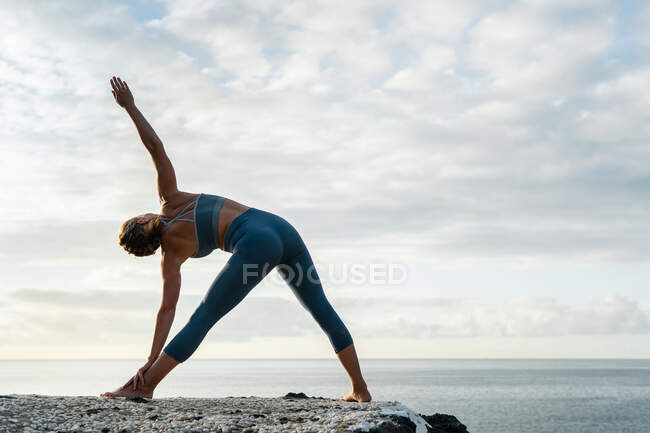 Задний вид неузнаваемой женщины в спортивной одежде, стоящей в позе Триконасаны на коврике для йоги против колеблющегося океана под облачным небом на закате — стоковое фото