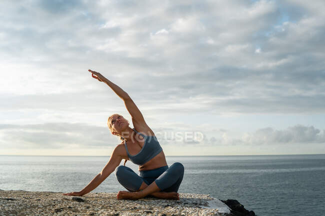 Young female in sportswear sitting with crossed legs while performing side bend and looking up on ocean coast in evening — Stock Photo