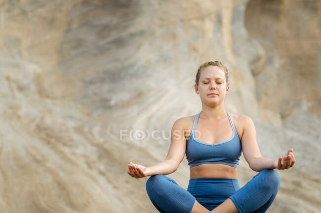 Joven mujer consciente en ropa deportiva meditando con los ojos cerrados mientras practica yoga sobre fondo rocoso - foto de stock