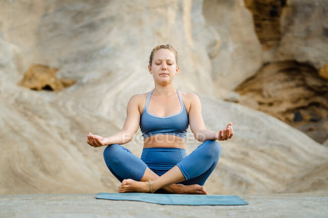 Молодая внимательная женщина в спортивной одежде медитирует с закрытыми глазами, практикуя йогу на скалистом фоне — стоковое фото
