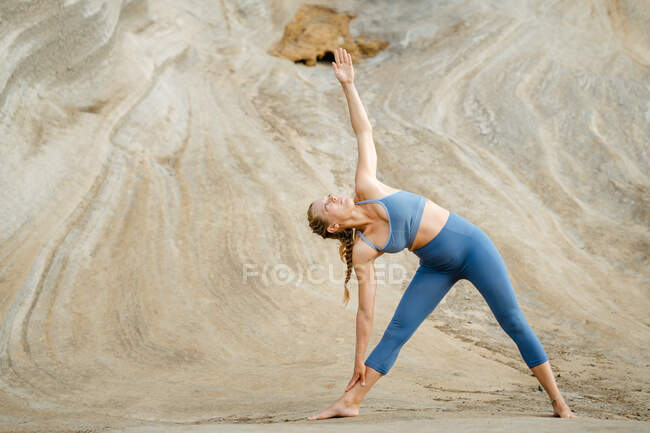 Молодая внимательная женщина в спортивной одежде в триконасане смотрит вверх, практикуя йогу на большом каменном фоне — стоковое фото
