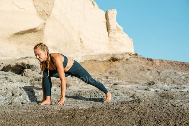 Молодая босая женщина в спортивной одежде, практикующая йогу на высоком выпаде позирует против скалистой горы при солнечном свете — стоковое фото