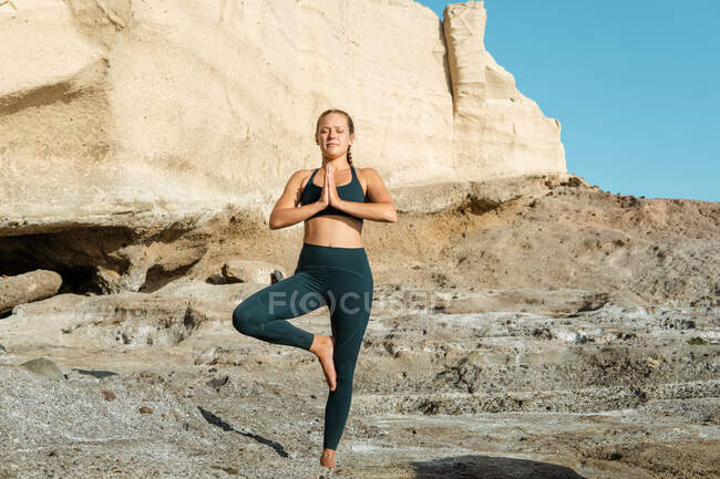 Joven mujer consciente en ropa deportiva en vrksasana con los ojos cerrados mientras practica yoga de pie sobre fondo rocoso - foto de stock