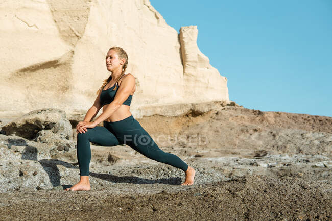 Young barefoot female in sportswear practicing yoga while touching knee and looking forward against mountain in sunlight — Stock Photo