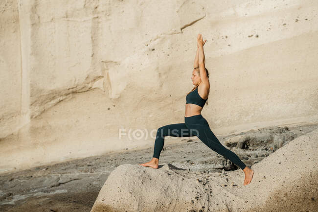 Jeune femme pieds nus en vêtements de sport pratiquant le yoga sur Crescent Pose contre la montagne rocheuse au soleil — Photo de stock