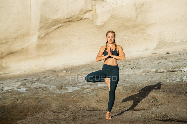Молода усвідомлена жінка в спортивному одязі на vrksana, дивлячись вниз, практикуючи йогу, що стоїть на кам'янистому фоні — стокове фото