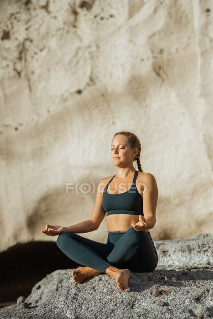 Jeune femme consciente en vêtements de sport méditant avec les yeux fermés tout en pratiquant le yoga assis sur un fond rocheux — Photo de stock
