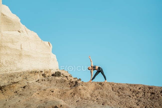 De baixo de jovem fêmea descalça em sportswear praticando ioga em pose trikonasana com os olhos fechados contra a montanha rochosa na luz solar — Fotografia de Stock