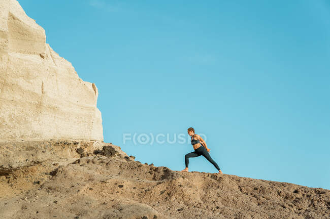 Desde abajo joven hembra descalza en ropa deportiva practicando yoga en la pose de Baddha Virabhadrasana con los ojos cerrados contra la montaña rocosa a la luz del sol - foto de stock