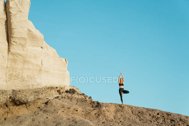 Joven mujer consciente en ropa deportiva en vrksasana con los ojos cerrados mientras practica yoga de pie sobre fondo rocoso - foto de stock