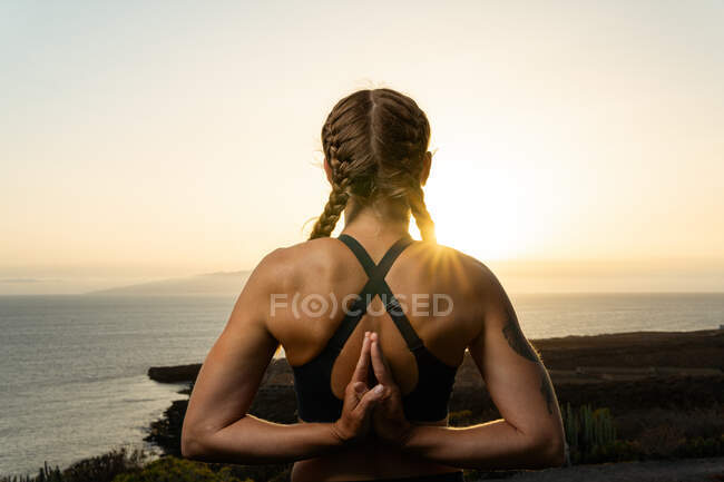 Vista posteriore di femmina anonima con mani namaste che praticano yoga contro l'oceano al crepuscolo nella retroilluminazione — Foto stock