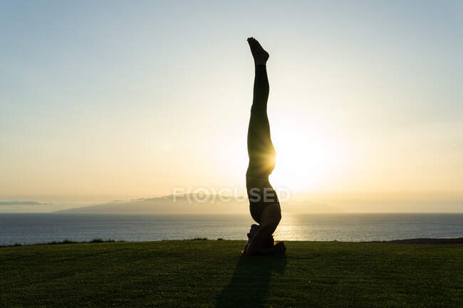 Vista lateral de la silueta femenina anónima de pie sobre la cabeza mientras practica yoga en la costa del océano a la luz del sol al atardecer - foto de stock