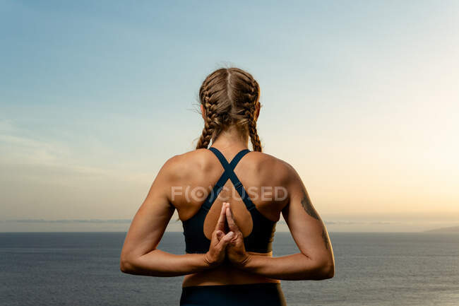 Vista posterior de una mujer anónima con manos namaste practicando yoga contra el océano en el crepúsculo con retroiluminación - foto de stock