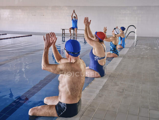 Gruppe von Menschen in Badebekleidung, die am Pool sitzen und die erhobenen Arme ausstrecken, während sie während des Wassergymnastik-Trainings mit dem Trainer im Pool trainieren — Stockfoto