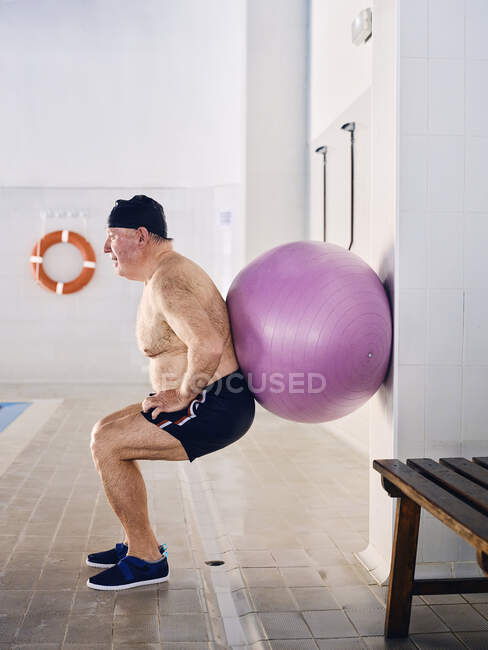 Vista lateral del nadador masculino de mediana edad haciendo ejercicios en cuclillas con balón en forma durante el entrenamiento aeróbico en la piscina - foto de stock
