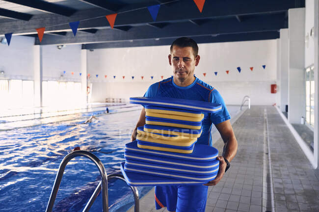 Seriöser männlicher Schwimmlehrer läuft nach dem Training mit einem Stapel Schaumstoffbrettern am Beckenrand entlang — Stockfoto