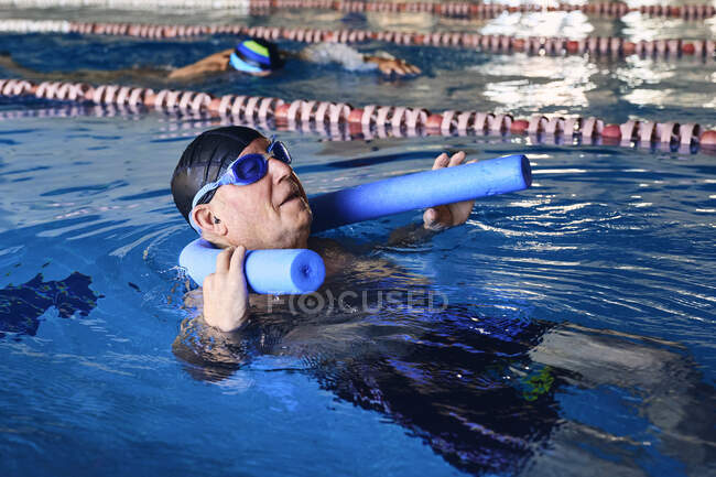 Вид со стороны мужчины средних лет в очках, плавающего на спине на пенной лапше во время водной аэробики в бассейне — стоковое фото