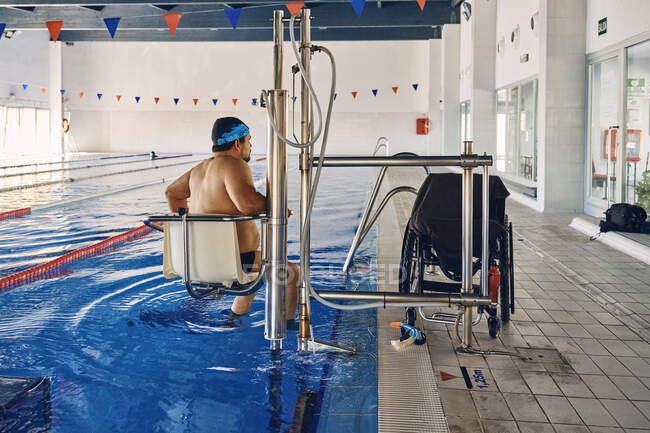 Homme mature assis sur la chaise de la piscine ascenseur près du fauteuil roulant avant l'entraînement — Photo de stock