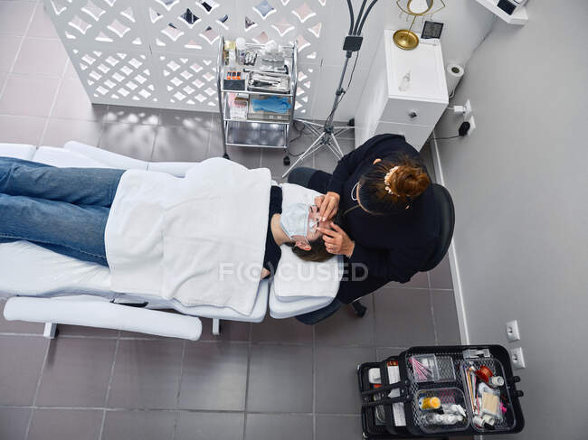 Сверху профессиональный косметолог наносит чистящее средство на ресницы клиентки во время процедуры красоты в салоне — стоковое фото