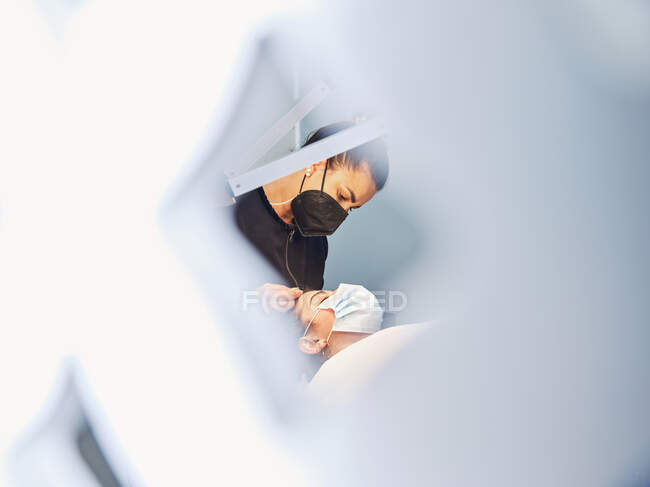 Através da vista lateral de grelha do profissional cosmetician que trata a cara do cliente feminino na máscara protetora durante o procedimento de beleza no salão — Fotografia de Stock