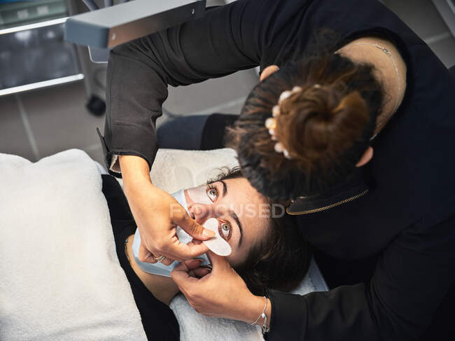 Сверху профессиональный косметик наносит пластыри под глазами клиентки во время процедуры наращивания ресниц в салоне — стоковое фото