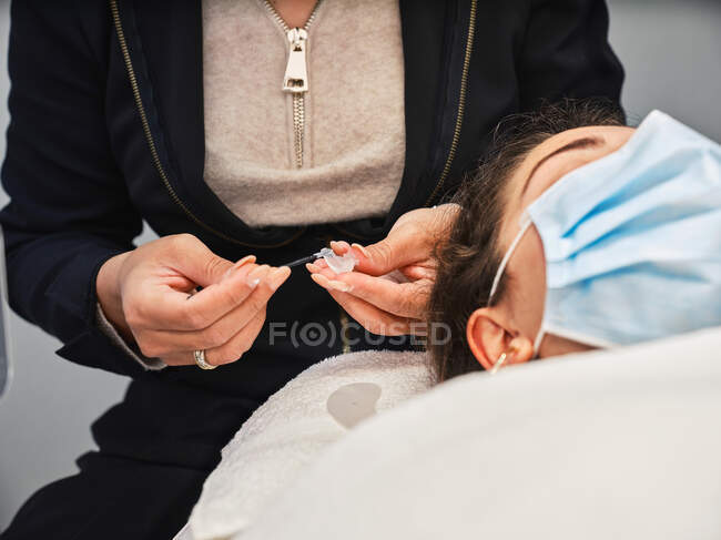 Crop cosmetico anonimo facendo procedura di estensione ciglia per il cliente femminile in maschera protettiva durante la sessione di bellezza nel salone — Foto stock