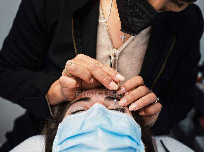 Close-up de cultura cosmetician anônimo com aplicador cobrindo pestanas de mulher com solução para levantamento de pestanas durante o procedimento de beleza no salão — Fotografia de Stock