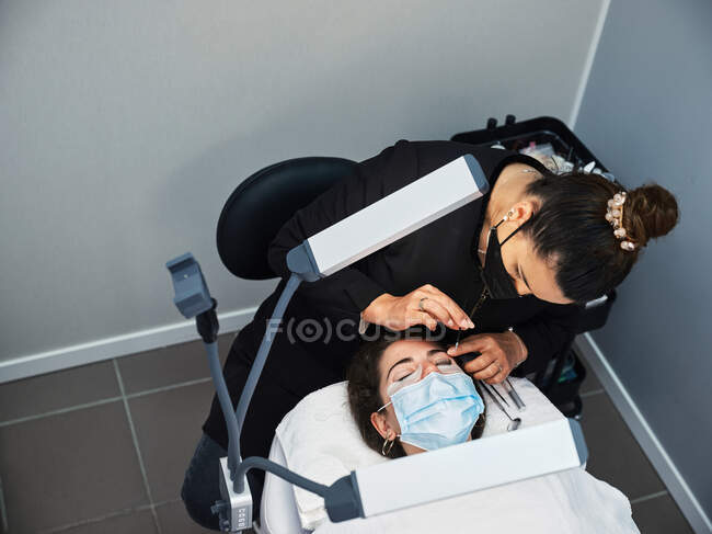 From above cosmetician with applicator covering eyelashes of woman with solution for lash lifting during beauty procedure in salon — Stock Photo