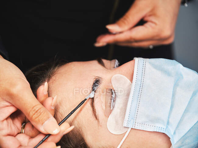 Primer plano de cultivo esteticista anónima con aplicador que cubre la ceja del cliente femenino con el pasado para la laminación durante el tratamiento de belleza en el salón - foto de stock