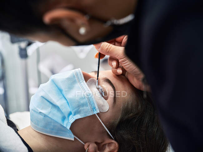 Visão lateral da cultura profissional cosmetician aplicação de solução em pestanas de paciente feminino em máscara protetora durante o procedimento de tratamento de beleza no salão — Fotografia de Stock