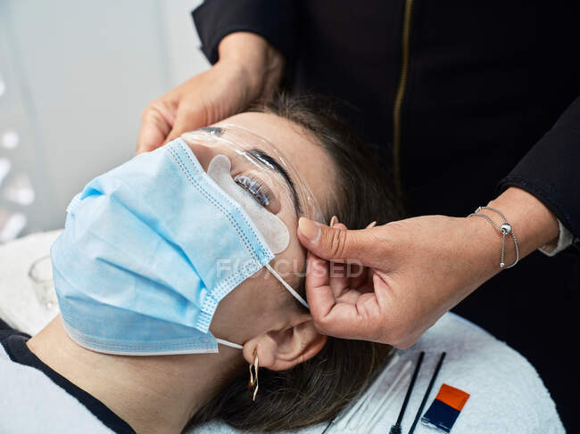 Crop anonymous cosmetician covering eyebrows of female client with plastic film during lamination procedure in beauty salon — Stock Photo