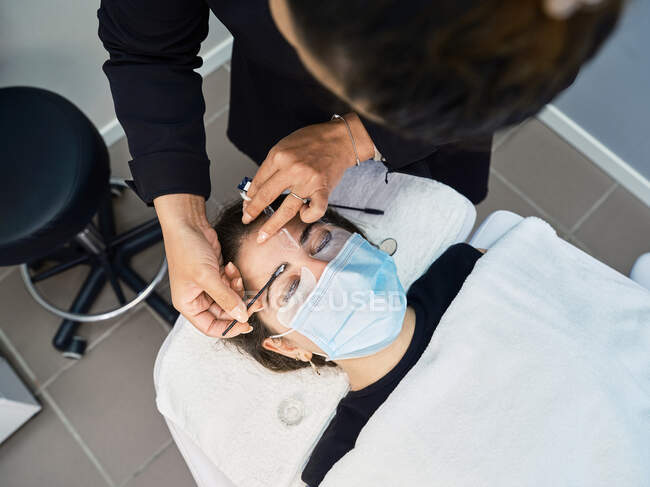 From above crop anonymous beautician with applicator covering eyebrow of female client with past for lamination during beauty treatment in salon — Stock Photo