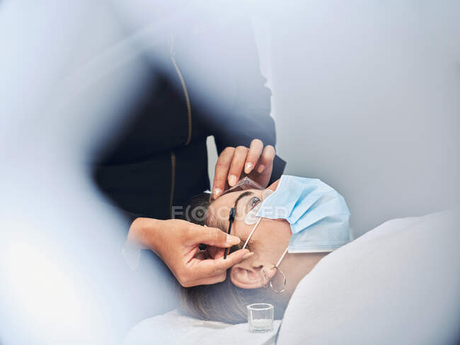 Crop anonymous beautician with applicator covering eyebrow of female client with past for lamination during beauty treatment in salon — Stock Photo