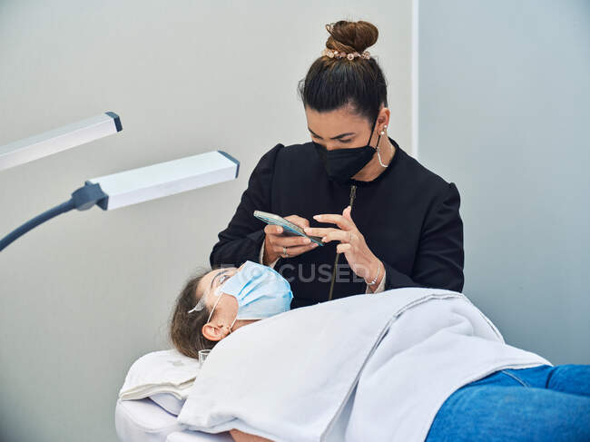 Cosmetologist profissional com smartphone tirando foto do rosto do cliente feminino recebendo tratamento de cílios durante o procedimento de beleza no salão — Fotografia de Stock