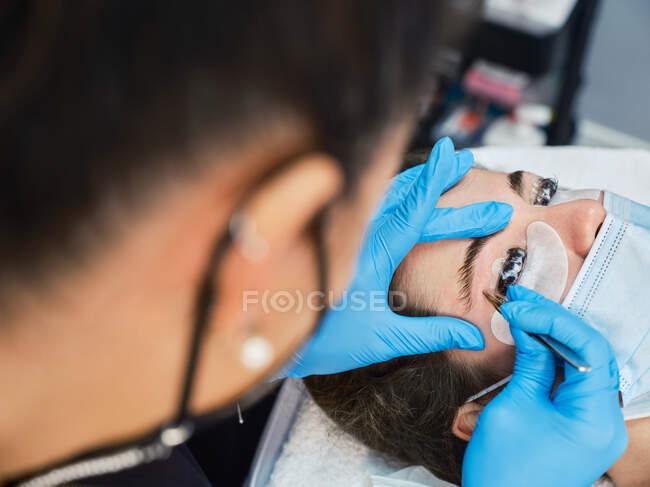 Cropped unrecognizable professional cosmetician in latex gloves using tweezers while modelling eyebrows of female patient during beauty procedure in salon — Stock Photo