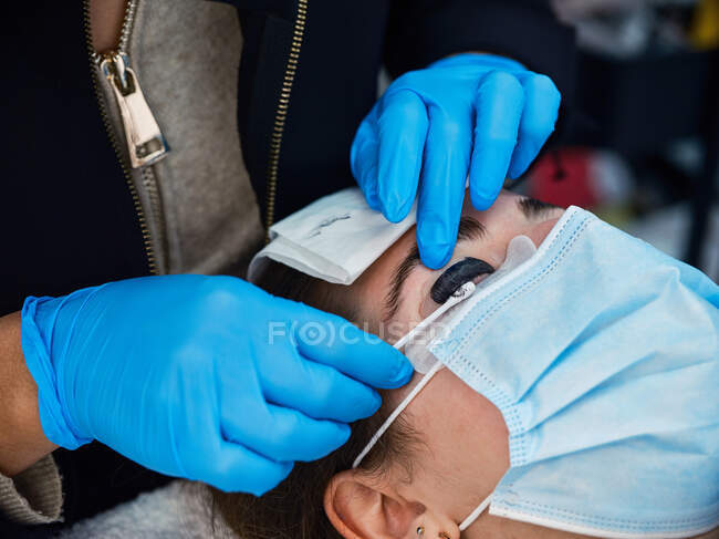 Crop cosmetologist in latex gloves with applicator treating eyelashes of female client during extension and lamination procedure in salon — Stock Photo
