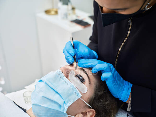 Cropped unrecognizable professional cosmetician in latex gloves using tweezers while modelling eyebrows of female patient during beauty procedure in salon — Stock Photo
