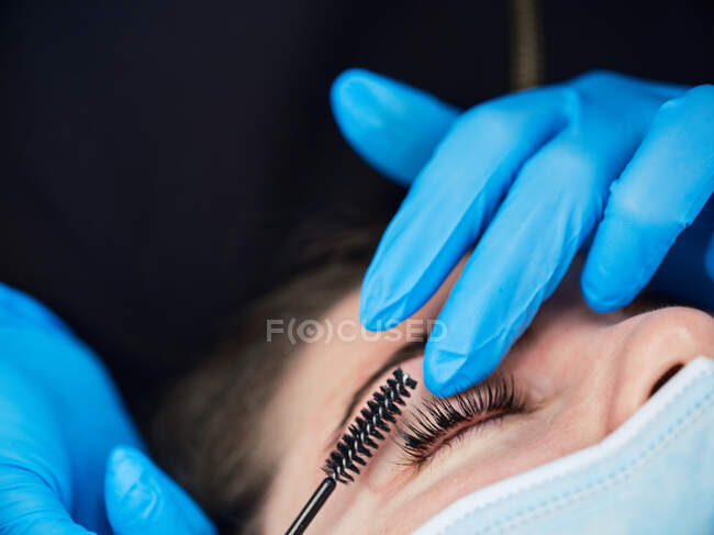 Closeup of crop anonymous cosmetologist in latex gloves using eyelash curler brush during beauty procedure to female customer — Stock Photo