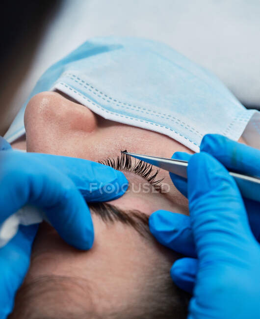 Closeup of crop anonymous beautician in latex gloves using tweezers while applying long eyelashes on eyelid of female client in salon — Stock Photo