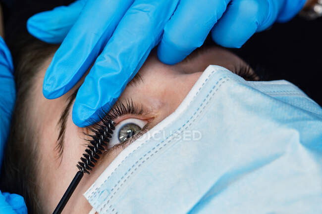 Nahaufnahme einer anonymen Kosmetikerin in Latex-Handschuhen mit Wimpernzange während eines Schönheitsprozesses für eine Kundin — Stockfoto