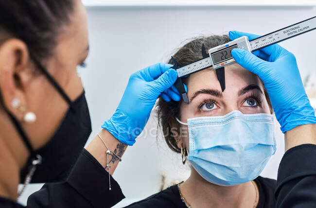 Вырезанный неузнаваемый косметолог в латексных перчатках с помощью линейки для измерения бровей клиента-женщины в защитной маске, получающей косметическую процедуру во время пандемии коронавируса — стоковое фото