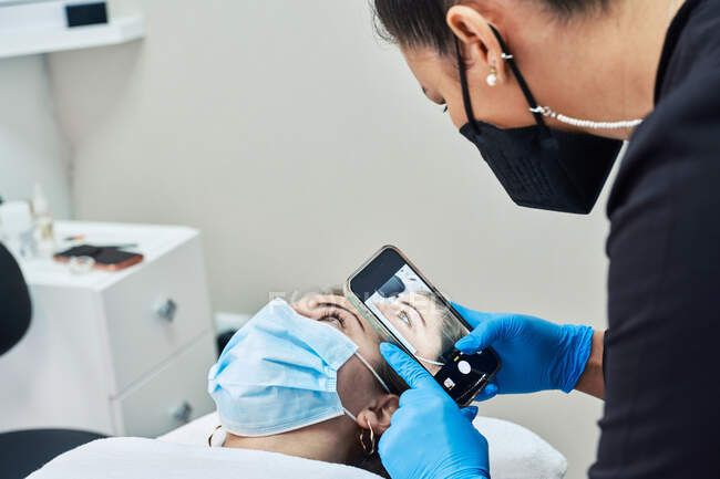 Профессиональный косметолог со смартфоном фотографирует лицо клиентки, получающей лечение ресниц во время процедуры красоты в салоне — стоковое фото