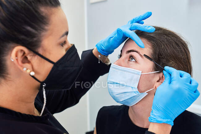 Esthéticienne en gants de latex brossant les sourcils d'une jeune cliente en masque de protection lors d'un rendez-vous beauté dans un salon moderne — Photo de stock